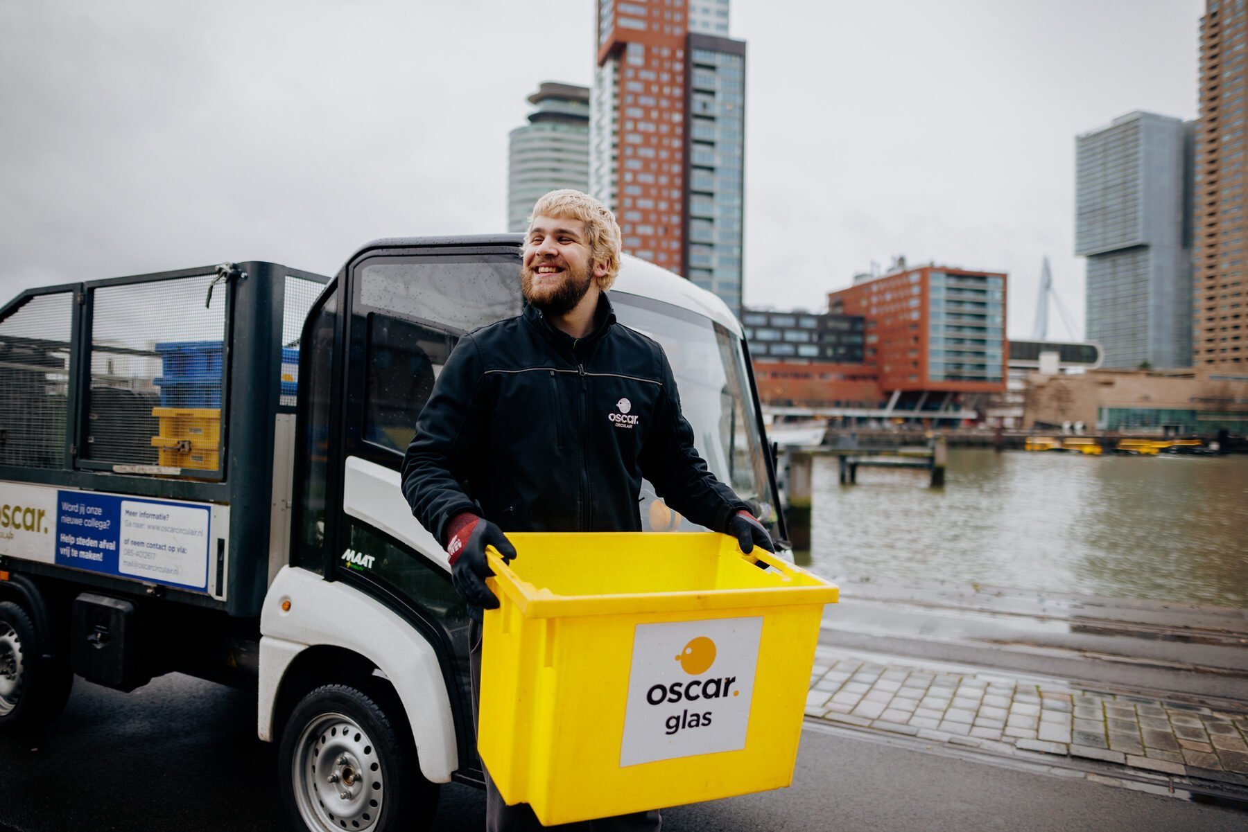 An Oscar Circulair collector carrying a yellow glass crate beside a light electric vehicle on the Rotterdam waterfront